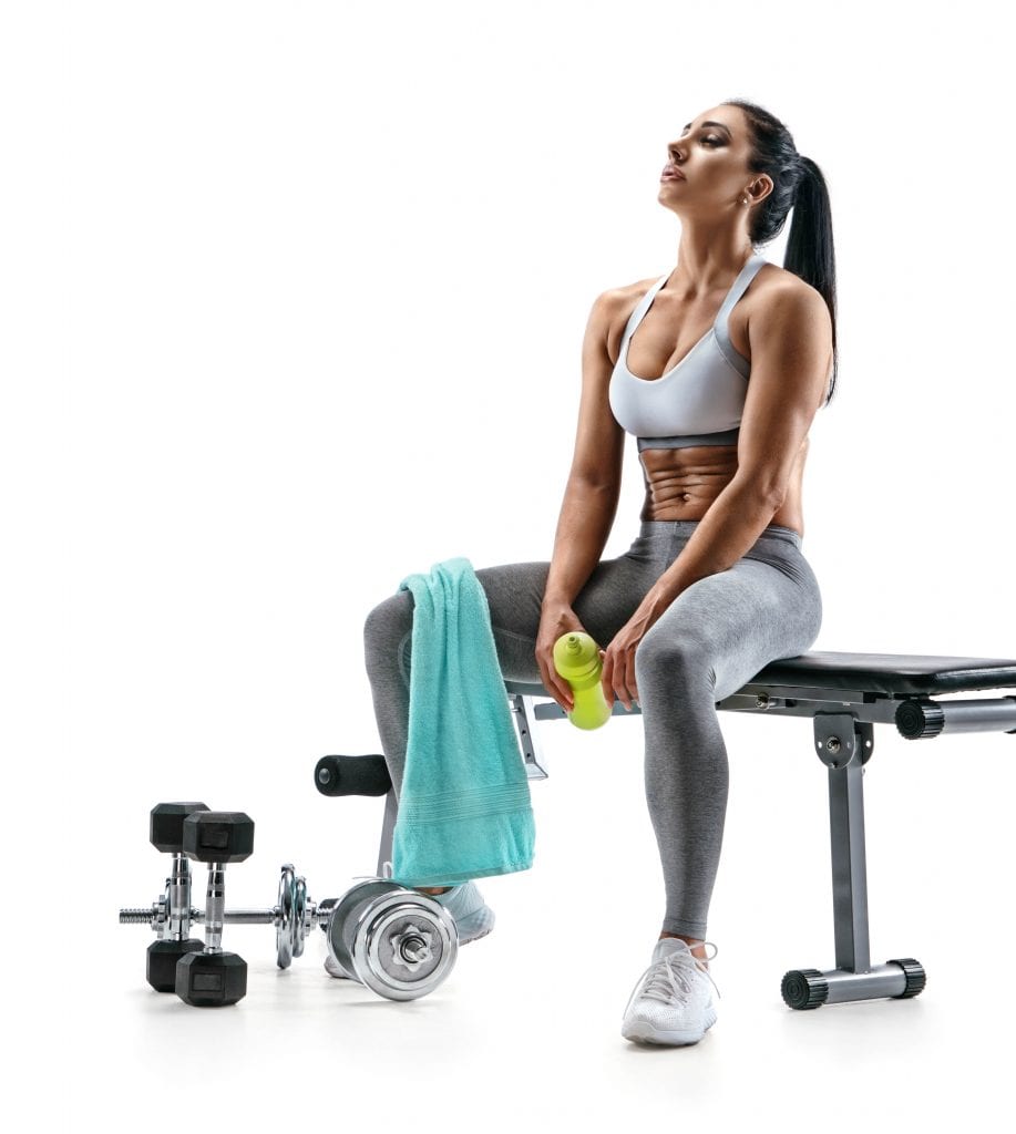 Femme d'origine latine assise sur un banc de musculation, avec une serviette sur la jambe droite et une gourde à la main. Des haltères jonchent le sol